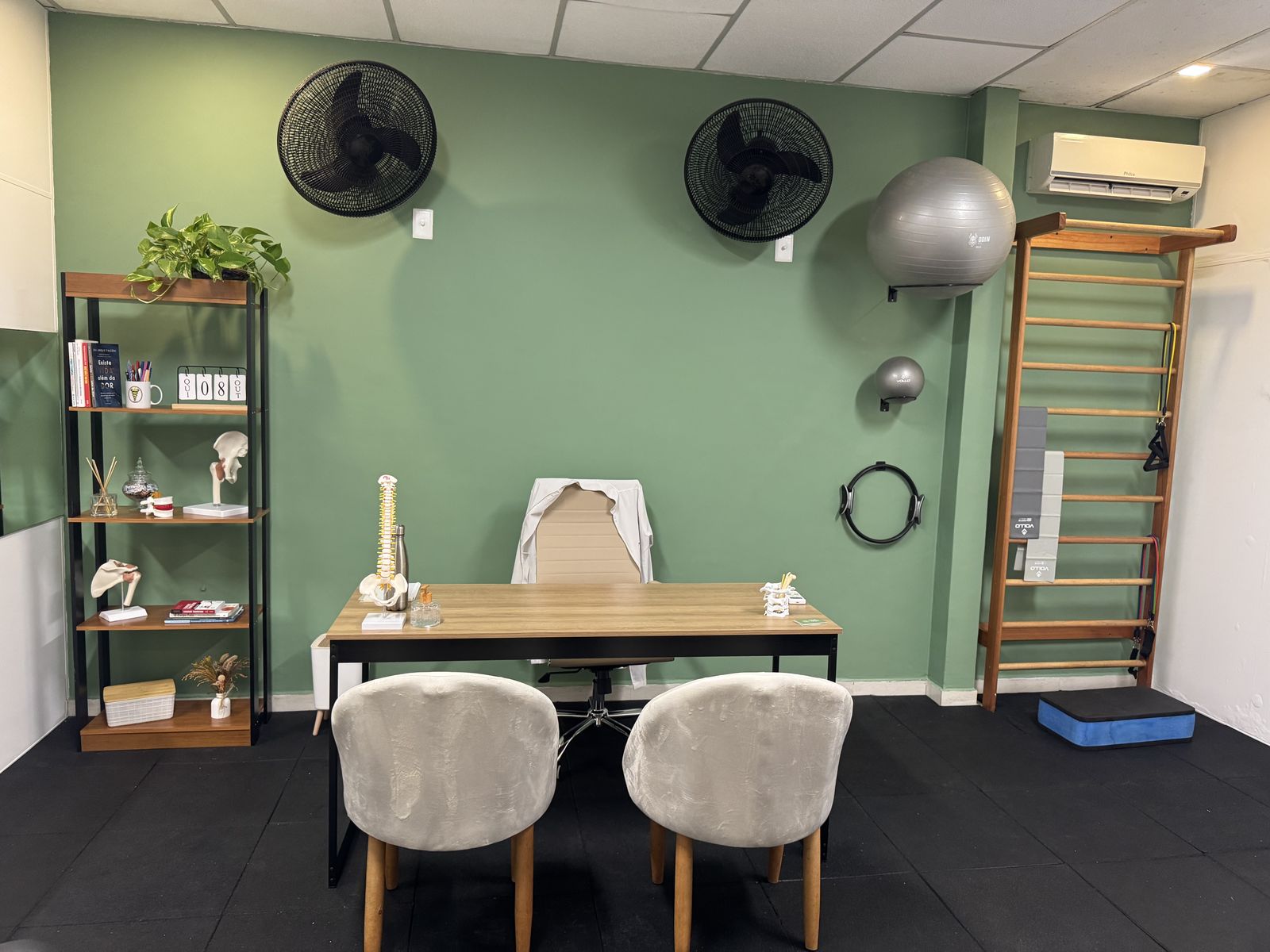 Mesa de avaliação, cadeiras e espaldar na parede verde do consultório de fisioterapia